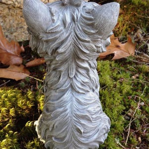 May include: A grey sculpture of a dog's head and back, seen from behind. The statue has detailed fur and ears, with a small decorative element on top. The dog statue is set against a backdrop of moss and leaves.
