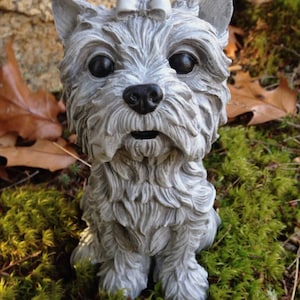 May include: A grey dog statue, possibly a Yorkshire Terrier, with a bow, black eyes and nose. The dog is sitting on green moss. The statue is outdoors, with a rock in the background.