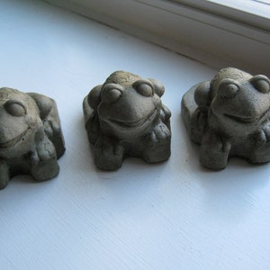 May include: Three small, gray concrete frog figurines with smiling faces and closed eyes. The frogs are in a row, each with a slightly different pose. The figurines are on a white surface.