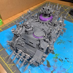 May include: A gray plastic model tank with numerous spikes, skulls, and a multi-barrel rocket launcher. The tank has a purple turret base and is detailed with various weapon attachments. The model is on a blue cutting mat.
