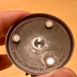 May include: A close-up of the underside of a dark gray, circular object. The object has three small, silver-colored magnets and several lines of text, including "Games Workshop Limited 2022" and a product number.