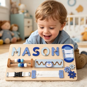 Könnte beinhalten: Holz-Aktivitätstafel mit dem Namen "MASON" in blauen Buchstaben. Die Tafel verfügt über interaktive Elemente wie einen Reißverschluss, Zahnräder, einen Pinguin und ein Perlenlabyrinth. Entwickelt für Kleinkinder.