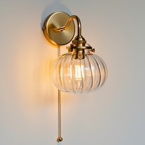 Könnte beinhalten: Eine Wandleuchte mit einem transparenten, gerippten Glasschirm und einem messingfarbenen Metallsockel. Die Leuchte hat eine Zugkette und eine Glühbirne im Vintage-Stil. Die Leuchte ist an einer weißen Wand montiert.