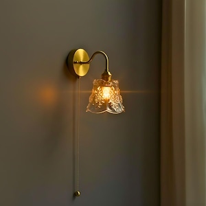 Può includere: Applique da parete in ottone con paralume in vetro trasparente a motivi floreali. L'apparecchio è montato su una parete grigia e ha una catena. L'applique emette una luce calda, ideale per l'illuminazione ambientale.