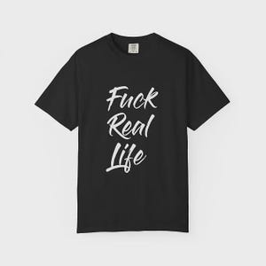 Könnte beinhalten: Schwarzes T-Shirt mit dem weißen Text "Fuck Real Life" in handschriftlichem Stil. Das Shirt hat kurze Ärmel und einen Rundhalsausschnitt. Der Text ist mittig auf der Vorderseite des Shirts platziert.