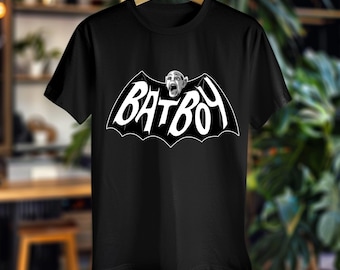 May include: Black t-shirt featuring a white graphic of the word "BATBOY" in a bat-wing shape, with a screaming face in the center. The shirt is hanging on a wooden hanger.