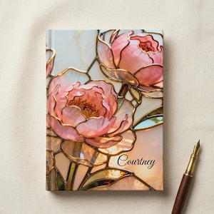 Op de afbeelding: Een gepersonaliseerd notitieboek met een glas-in-lood bloemenontwerp met roze en perzik pioenrozen. De kaft heeft een gouden omtrek en de naam "Courtney" in zwart schrift. Een bruine en gouden pen ligt ernaast.