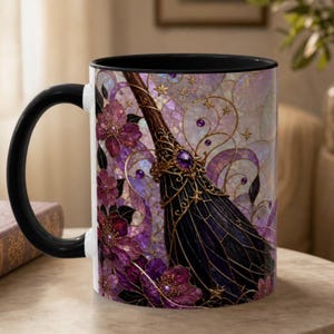 May include: A ceramic mug with a black handle and interior, featuring a stained-glass style design of a dark broomstick adorned with gold filigree and a purple gemstone, surrounded by purple and pink flowers, swirling vines, and small gold stars.