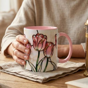 K&ouml;nnte beinhalten: Eine wei&szlig;e Keramiktasse mit rosa Griff und Innenraum, verziert mit einem Tulpenmuster im Glasmalerei-Stil in Rosa- und Gr&uuml;nt&ouml;nen. Die Tasse wird von einer Person gehalten.