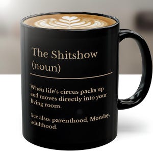 Puede incluir: Taza de cerámica negra con el texto "The Shitshow (noun)" en una fuente serif. La taza contiene un café con leche con un diseño en forma de corazón. El texto adicional incluye "When life's circus packs up and moves directly into your living room."