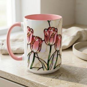 K&ouml;nnte beinhalten: Eine wei&szlig;e Keramiktasse mit rosa Innenraum und Henkel. Die Tasse zeigt ein Buntglasdesign aus rosa Tulpen mit goldenen Akzenten und gr&uuml;nen Stielen. Die Tasse steht auf einer hellen Oberfl&auml;che.