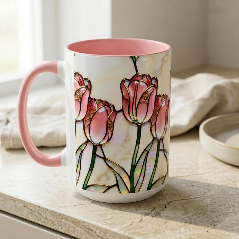 K&ouml;nnte beinhalten: Eine wei&szlig;e Keramiktasse mit rosa Innenraum und Henkel. Die Tasse zeigt ein Buntglasdesign aus rosa Tulpen mit goldenen Akzenten und gr&uuml;nen Stielen. Die Tasse steht auf einer hellen Oberfl&auml;che.