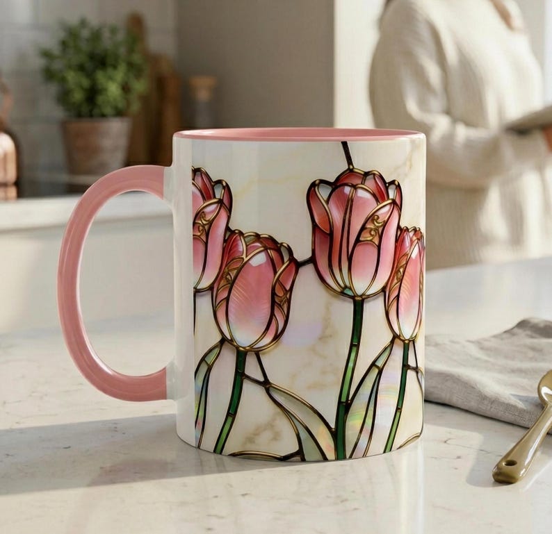 K&ouml;nnte beinhalten: Eine Keramiktasse mit rosa Griff und Rand, mit einem Buntglasdesign aus rosa Tulpen mit goldenen Umrissen. Der Hintergrund ist cremefarben. Die Tasse steht auf einer wei&szlig;en Oberfl&auml;che.