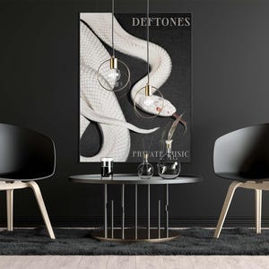Op de afbeelding: Een zwart-wit poster met een gestileerde slang en de bandnaam "Deftones". De poster hangt aan een zwarte muur, met twee zwarte stoelen en een ronde tafel ervoor.
