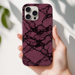 Puede incluir: Funda de teléfono burdeos con un diseño de encaje negro. La funda tiene un acabado mate y un recorte para la cámara para una cámara de múltiples lentes. La funda se sostiene en una mano sobre un fondo blanco.