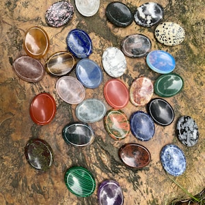 May include: A collection of oval-shaped gemstones in various colors and patterns, including red, blue, green, and white. The stones are arranged on a textured, brown surface, showcasing their polished surfaces and natural variations.