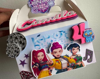 May include: White party favor box decorated with K-Pop themed stickers. The box features illustrations of three female characters, a disco ball, stars, a diamond, and the number 4. The box also has the name "Camila" and the word "HUNTRX".