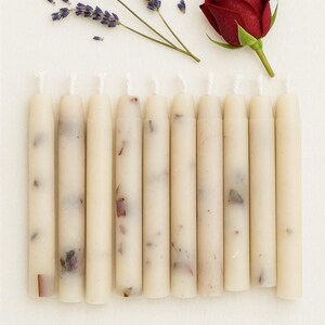 May include: A collection of ten cream-colored, slender candles, each adorned with embedded floral elements. A sprig of lavender and a deep red rose are arranged nearby, creating a serene and natural aesthetic.
