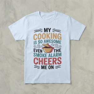 Puede incluir: Camiseta blanca con el texto "MY COOKING IS SO AWESOME EVEN THE SMOKE ALARM CHEERS ME ON" en varios colores. El diseño incluye una olla de dibujos animados.