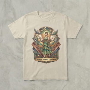 Puede incluir: Camiseta color crema con una ilustración detallada de una mujer con un vestido verde sosteniendo bolsas de compras. El diseño incluye elementos art déco, pájaros y el texto "Shopping is always a good idea."