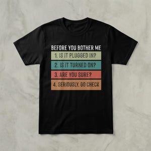 May include: Black t-shirt with the text "BEFORE YOU BOTHER ME" above a list of troubleshooting steps: "1. IS IT PLUGGED IN?", "2. IS IT TURNED ON?", "3. ARE YOU SURE?", and "4. SERIOUSLY, GO CHECK". The text is in white, green, blue, red, and orange.