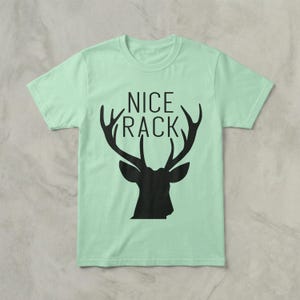 Puede incluir: Camiseta verde claro con las palabras "NICE RACK" sobre una silueta negra de la cabeza y cuernos de un ciervo. La camiseta es de cuello redondo y parece estar hecha de algodón o un tejido similar.