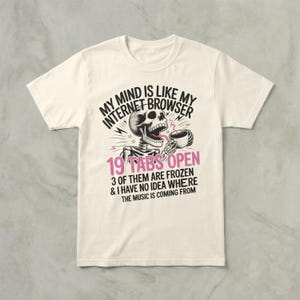 May include: Cream-colored t-shirt with a humorous graphic. The design features a skeleton holding a cup, with text that reads "MY MIND IS LIKE MY INTERNET BROWSER 19 TABS OPEN." Additional text states "3 OF THEM ARE FROZEN & I HAVE NO IDEA WHERE THE MUSIC IS COMING FROM."