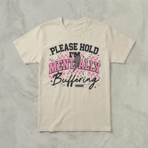 Puede incluir: Camiseta color crema con el texto "PLEASE HOLD I'M MENTALLY BUFFERING" en negro, rosa y blanco. El diseño incluye un rayo, margaritas y una barra de carga. La camiseta es de manga corta.