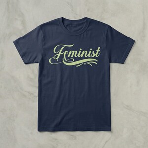 May include: Navy blue t-shirt with the word "Feminist" in a light green, cursive font. The word is centered on the shirt, with decorative flourishes above and below. The shirt is made of a soft material.