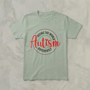 May include: A light green t-shirt with a circular graphic that reads "Seeing the World Autism Differently." The word "Autism" is in red script, surrounded by a dotted blue circle, with yellow and green accents.