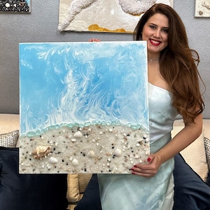 Puede incluir: Cuadro cuadrado de resina que representa una escena de playa. La mitad superior presenta un diseño azul y blanco arremolinado, que se asemeja a las olas del océano. La mitad inferior muestra una playa arenosa con conchas y pequeños guijarros. Decoración costera.