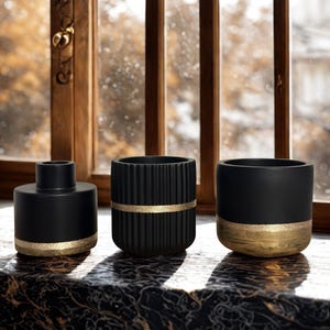 Könnte beinhalten: Drei schwarze Keramikvasen mit goldenen Akzenten stehen auf einer dunklen Oberfläche in der Nähe eines Fensters. Die Vasen haben unterschiedliche Formen: eine zylindrische Vase, eine geriffelte Vase und eine schalenförmige Vase. Die goldenen Bänder und Basen verleihen Eleganz.