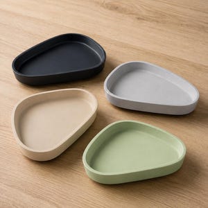 Peut inclure: Quatre petits plateaux peu profonds en forme de galet avec des contours triangulaires arrondis. L'ensemble comprend un plateau noir, un gris clair, un beige et un vert sauge, chacun avec une finition mate lisse et des bords surélevés.