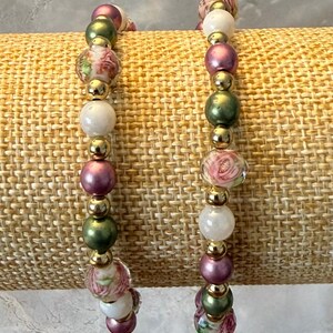 May include: Two beaded bracelets featuring a repeating pattern of round beads in metallic gold, olive green, and mauve, interspersed with white beads and clear glass beads decorated with pink floral rose patterns throughout the design.