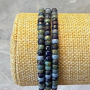 May include: Three stacked bracelets made of small, round, glazed ceramic beads in varying shades of blue, green, and dark purple. The beads feature a glossy, mottled finish with subtle color variations, arranged in a vertical column pattern.