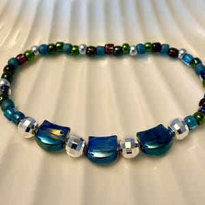May include: A beaded bracelet featuring three metallic blue cat-shaped beads interspersed with silver faceted spherical beads. The remainder of the bracelet consists of small, round glass beads in shades of green, blue, and silver with a glossy finish.