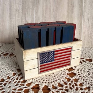 May include: A wooden box with a distressed white finish, featuring an embroidered American flag. Inside, there are several blue and red wooden blocks. The box sits on a white lace doily.