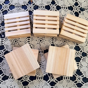 May include: A collection of light-colored wooden coasters, with three designed as miniature shipping pallets featuring horizontal slats and two featuring solid, smooth square surfaces, all showcasing a natural wood grain finish.
