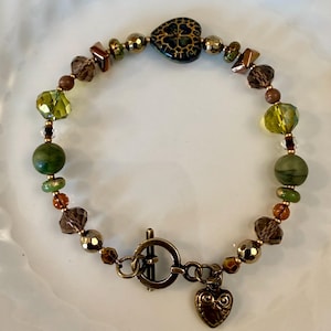 May include: A beaded bracelet featuring a mix of green, brown, and metallic beads, a central heart-shaped bead with a dark floral pattern, and a bronze-toned toggle clasp with a small heart-shaped charm dangling from the closure.