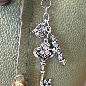 May include: A silver-toned metal bag charm featuring a decorative skeleton key pendant encrusted with clear rhinestones, a small heart-shaped charm with an embossed pattern, and a cylindrical bead charm accented with multiple clear rhinestones.