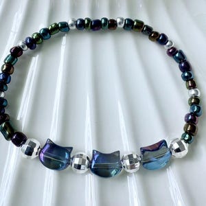 May include: A beaded bracelet featuring a cat-themed design. The bracelet incorporates iridescent cat-shaped beads, silver disco ball beads, and small, colorful beads in shades of blue, green, and purple. The bracelet is designed to be worn around the wrist.