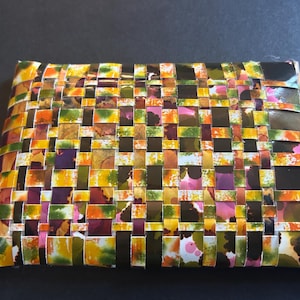 May include: A rectangular woven item with a colorful pattern. The design features interwoven strips in shades of yellow, orange, green, purple, and pink. The item appears to be made of a flexible material, possibly paper or fabric.