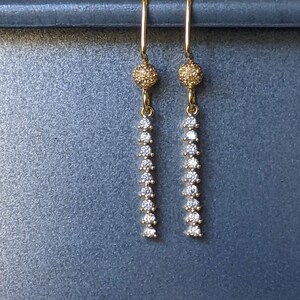 May include: A pair of gold-colored drop earrings. Each earring features a textured ball at the top, followed by a vertical line of small, clear gemstones. The earrings are displayed against a muted blue-gray background.