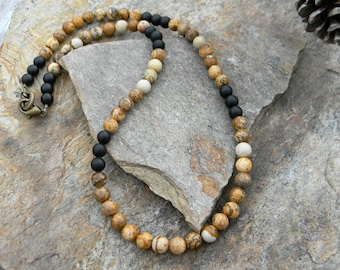 May include: A beaded necklace featuring a mix of natural stone beads in shades of tan, brown, and cream, interspersed with black beads. The necklace has a bronze-colored clasp and is displayed on a stone surface.