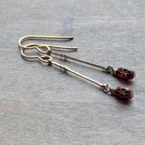 May include: A pair of gold-colored dangle earrings. Each earring features a long, slender bar with a decorative element and a small, red, oval-shaped bead with a starburst design. The earrings have a hook closure.