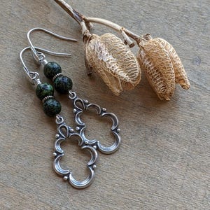 May include: A pair of silver earrings with a green and black speckled stone dangle. The earrings have a delicate, ornate design.