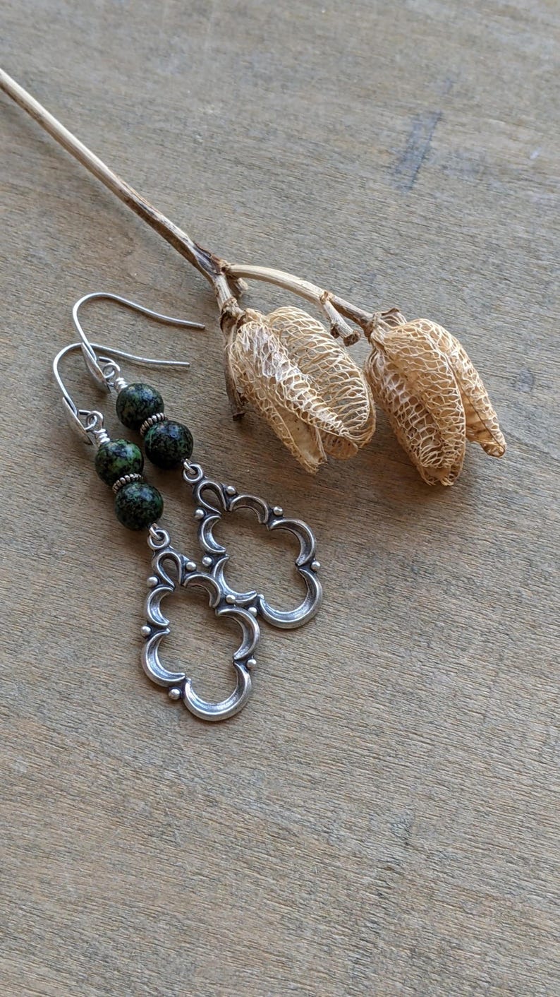 May include: A pair of silver earrings with a green and black speckled stone dangle. The earrings have a delicate, ornate design.