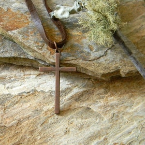 May include: A simple copper cross pendant on a brown leather cord. The pendant is hanging on a stack of brown rocks.