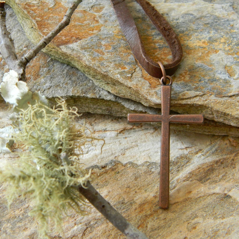 May include: A brown leather cord necklace with a copper cross pendant. The necklace is displayed on a background of stacked brown and gray stones.