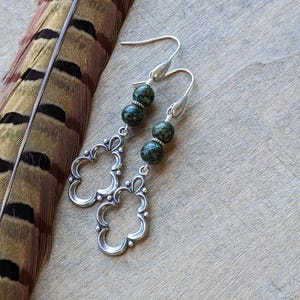 May include: Silver dangle earrings with two green and black speckled beads on each earring. The earrings have a floral design.